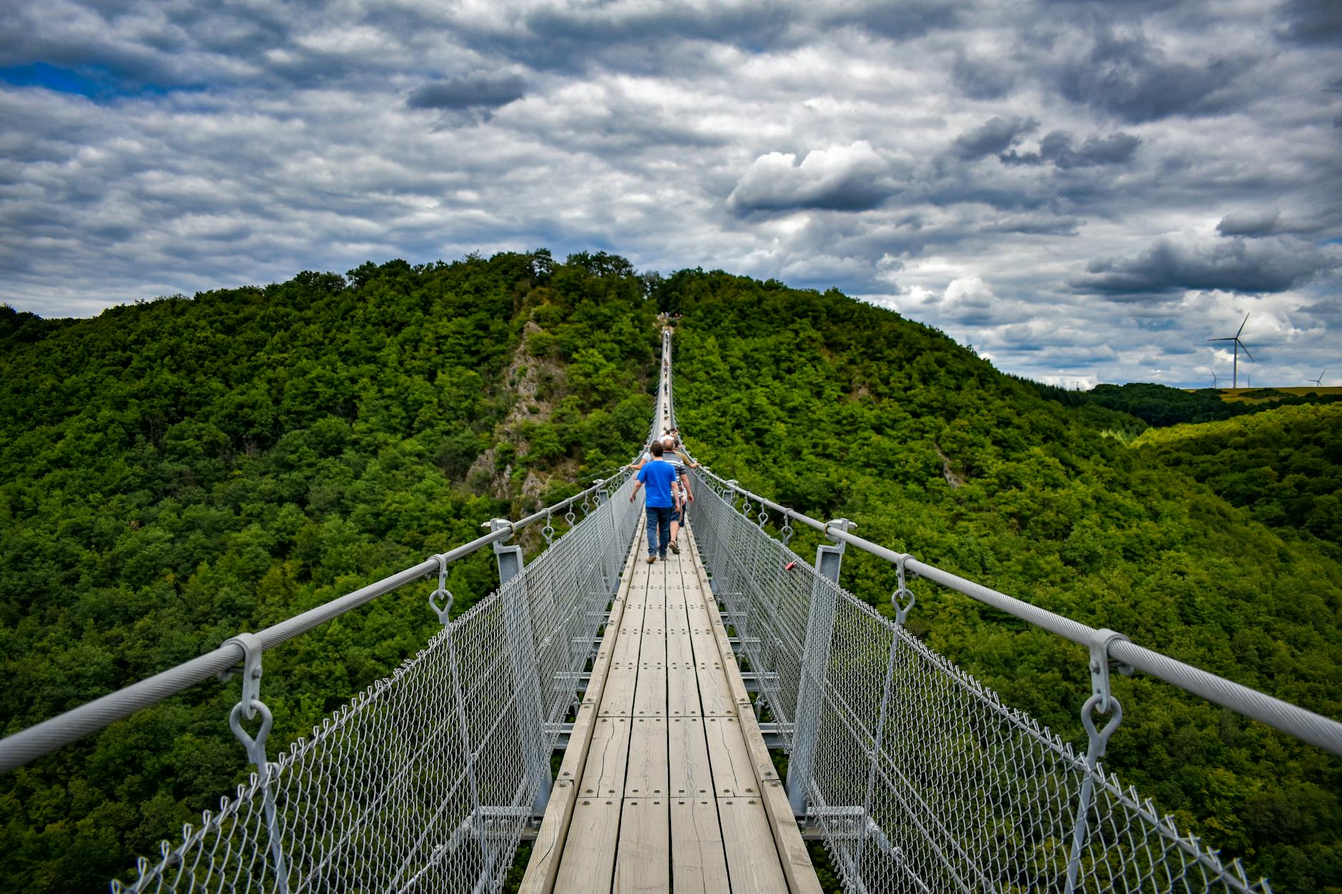 Tourists walking on the Geierlay Suspension Bridge surrounded by lush greenery and dramatic clouds.